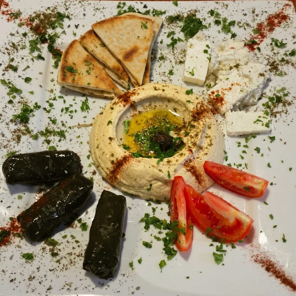 A white plate with hummus, pita bread, feta cheese, stuffed grape leaves, and tomato wedges.